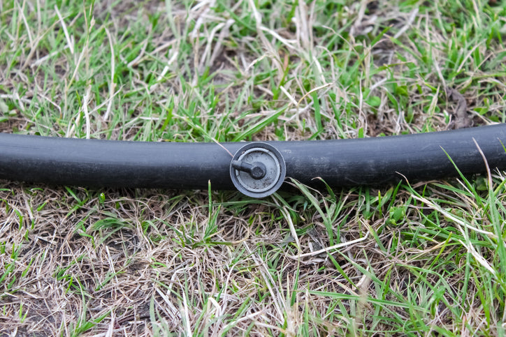 Close-up of an adjustable drip irrigation emitter installed on a flexible black plastic hose layout over green and dry grass in a backyard lawn